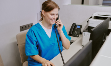 Young Adult Woman Answering Phone Call While Working at Reception Desk | Featured Image for the How to Manage Busy Clinic Phone Lines blog