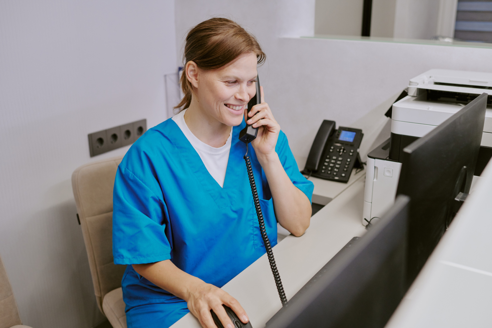 Young Adult Woman Answering Phone Call While Working at Reception Desk | Featured Image for the How to Manage Busy Clinic Phone Lines blog